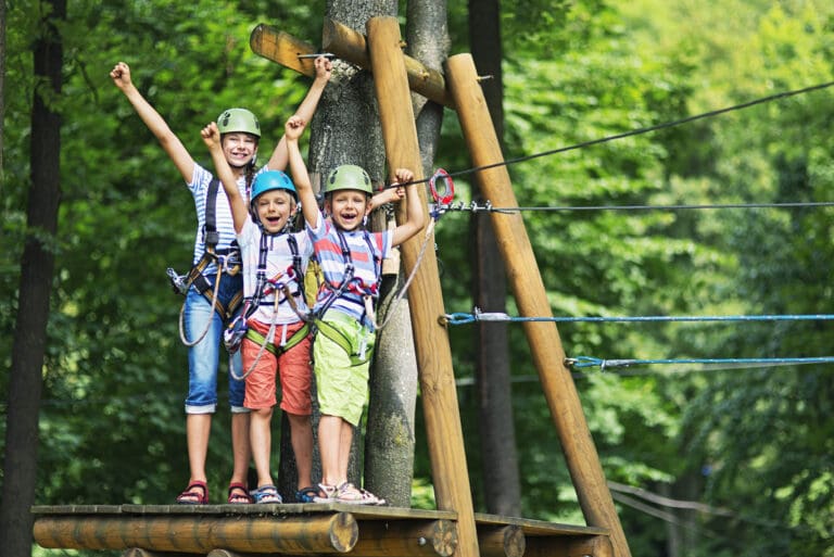 Little girl aged 10 with her brothers aged 7, wearing helmets stadning on wooden platform holding zip line in the outdoors ropes course adventure park. Kids are smiling at the camera and cheering.